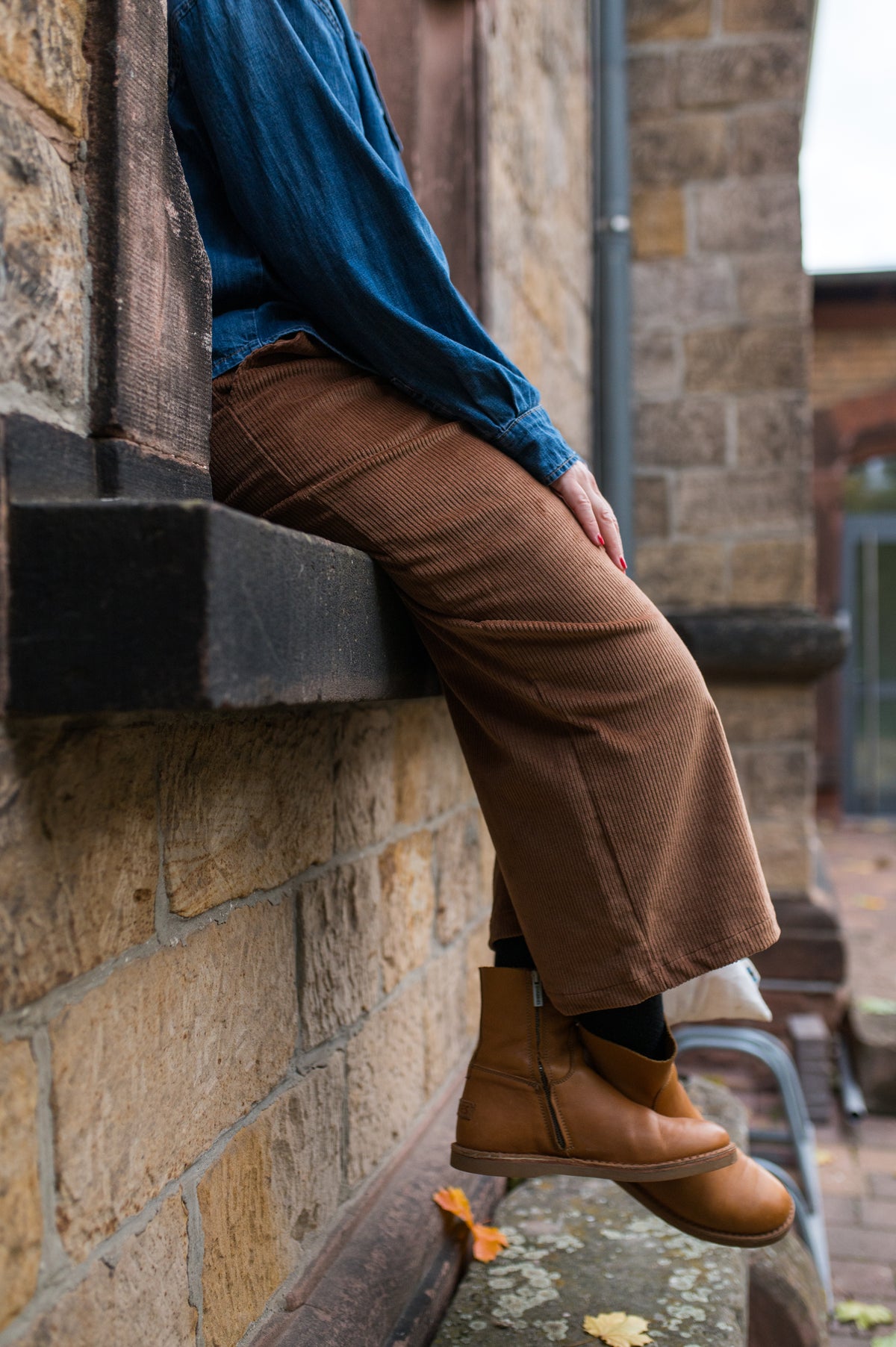 Frau sitzt auf einer Mauer in cognacfarbener Cord-Culotte, blauem Pulli und Stiefeln