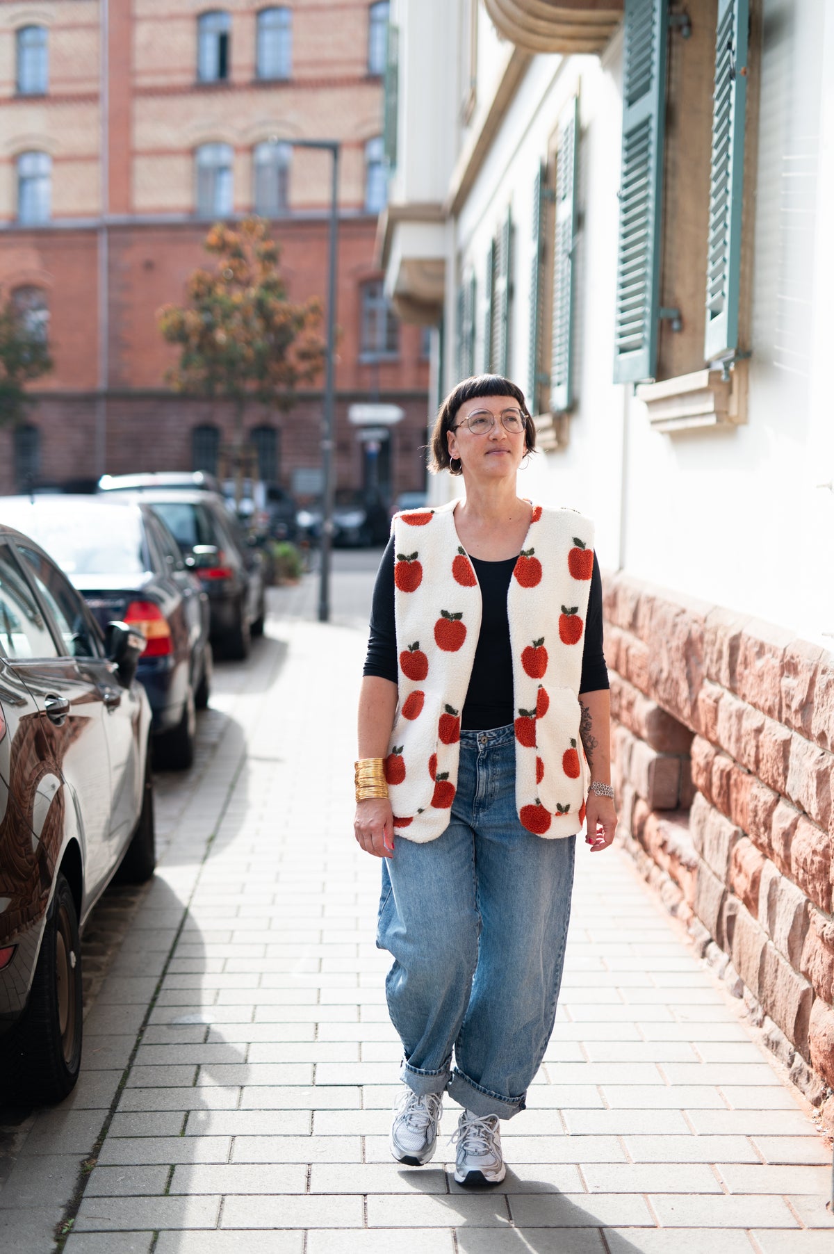 Frau in cremeweißer Oversize-Teddyweste mit roten Äpfeln, schwarzes Shirt und weite Jeans, auf einer Straße