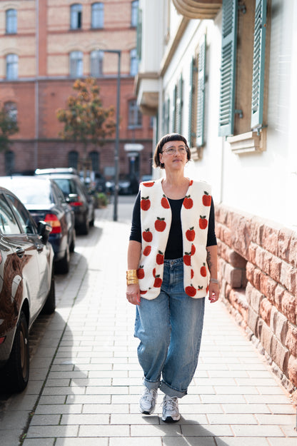 Frau in cremeweißer Oversize-Teddyweste mit roten Äpfeln, schwarzes Shirt und weite Jeans, auf einer Straße