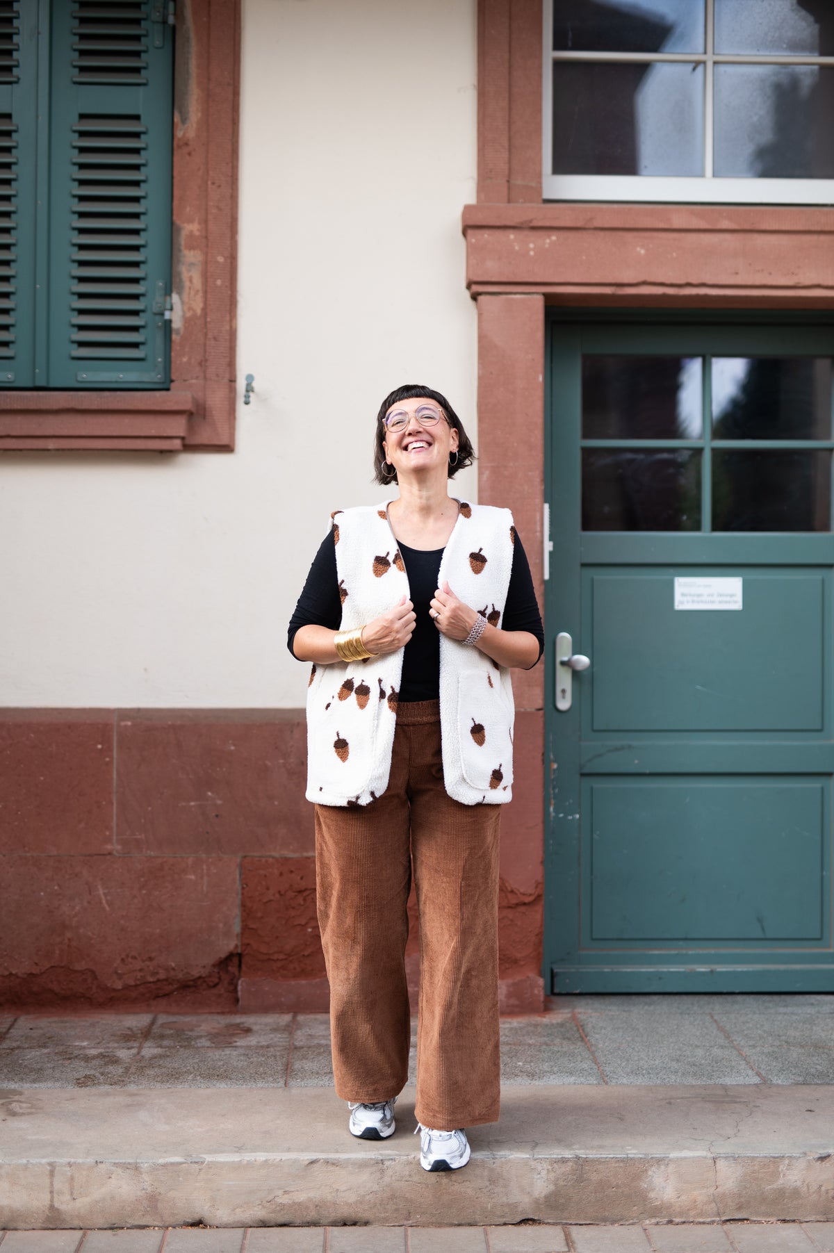 Lachende Frau in cremefarbener Oversize-Teddyweste mit braunen Eicheln, schwarzes Shirt und cognacfarbene Cordhose vor grüner Tür