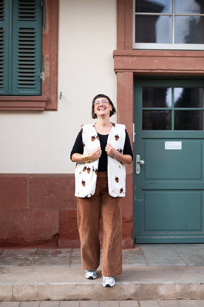 Lachende Frau in cremefarbener Oversize-Teddyweste mit braunen Eicheln, schwarzes Shirt und cognacfarbene Cordhose vor grüner Tür
