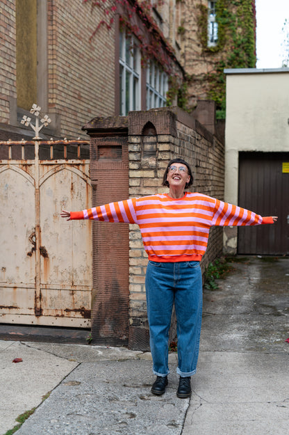 Frau mit ausgebreiteten Armen im orange-rosa gestreiften Oversize-Pullover mit weitem Ärmel