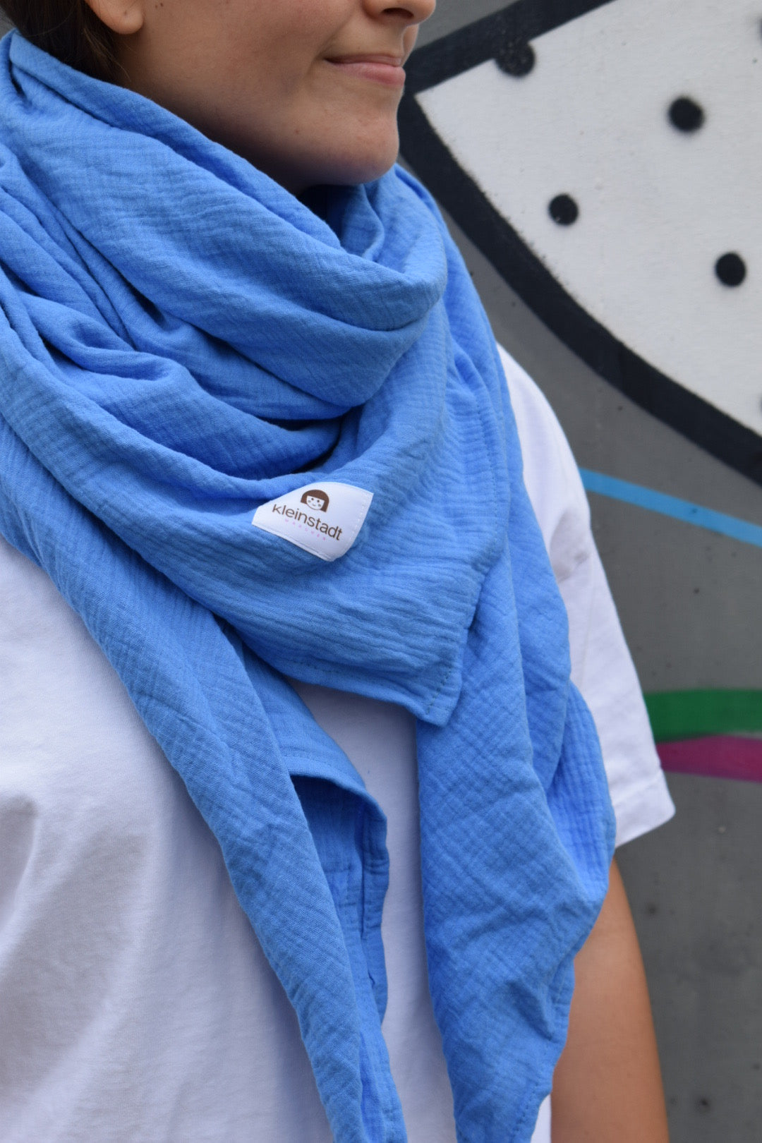 Frau trägt kornblumenblauen XXL-Musselinschal aus Bio-Baumwolle zu weißem Shirt vor Graffiti-Wand